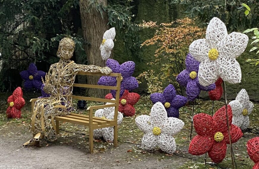 A wicker sculpture of a person sits on a bench, surrounded by large woven flowers in festive red, purple, and white hues, echoing the cheerful spirit of Jingle Bells in a garden with trees in the background.