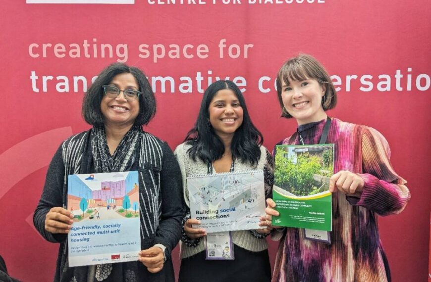 Three people smiling, each holding different posters, stand before a red backdrop emblazoned with "creating space for transformative conversation." The scene buzzes with energy reminiscent of Housing Central 2024 and captures the spirit of the Hey Neighbour Collective.
