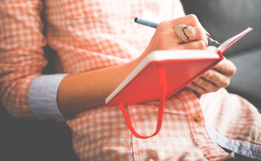 A person in a checkered shirt is journaling thoughtfully in a red notebook, enhancing their emotional intelligence as they sit.