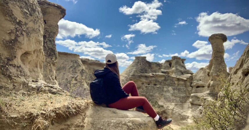 Person in a black jacket and red pants sits on a rock outside their comfort zones, overlooking a rugged desert landscape with a cloudy blue sky.