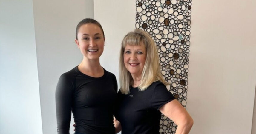 Two women in black shirts stand together, smiling proudly in front of a decorative wall panel, as if celebrating a unique patient graduation day.