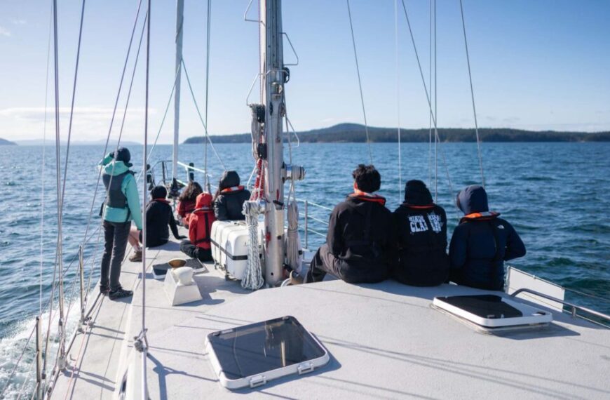 A group of people sits and stands on a sailboat, embracing the spirit of sailing in open water. The clear sky and distant land create a serene backdrop as they practice good stewardship of their journey.