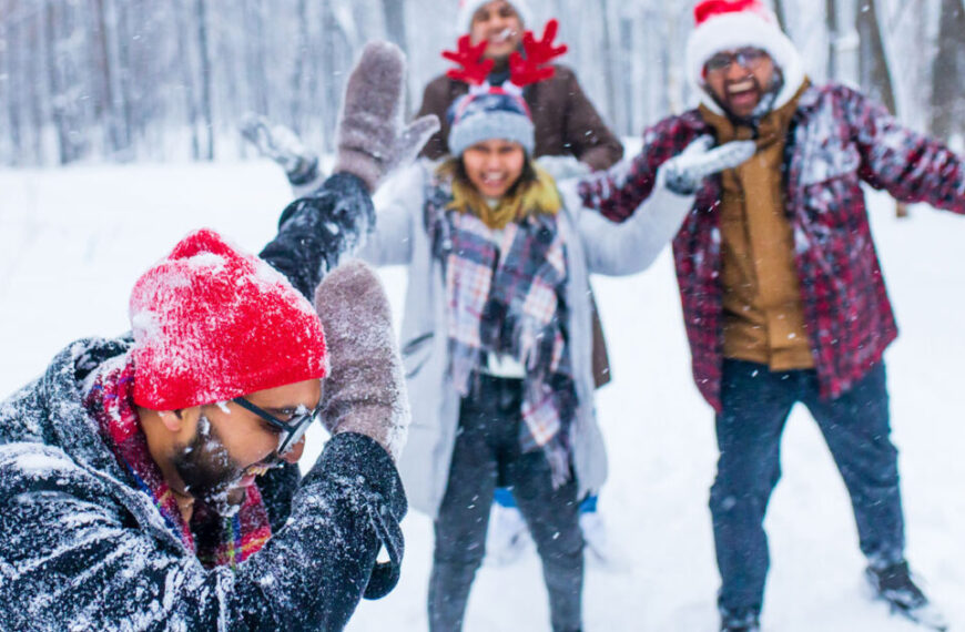Four people dressed in winter clothing play and laugh in a snowy forest, some wearing Santa hats and reindeer antlers, embracing the festive spirit of the holidays.