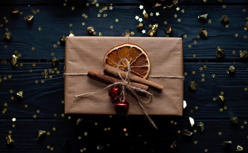 A gift wrapped in brown paper with a dried orange slice, cinnamon sticks, and a red bell on top captures the spirit of the giving season. Gold stars scattered around on a dark surface illuminate how small gestures can change lives.