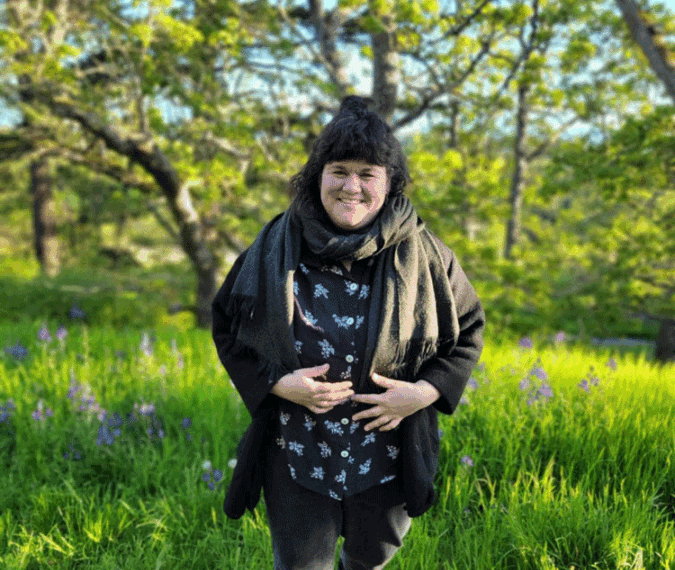 JO walks through a grassy area with trees in the background, wearing a black coat and dark pants, taking a pause to smile warmly at the camera.