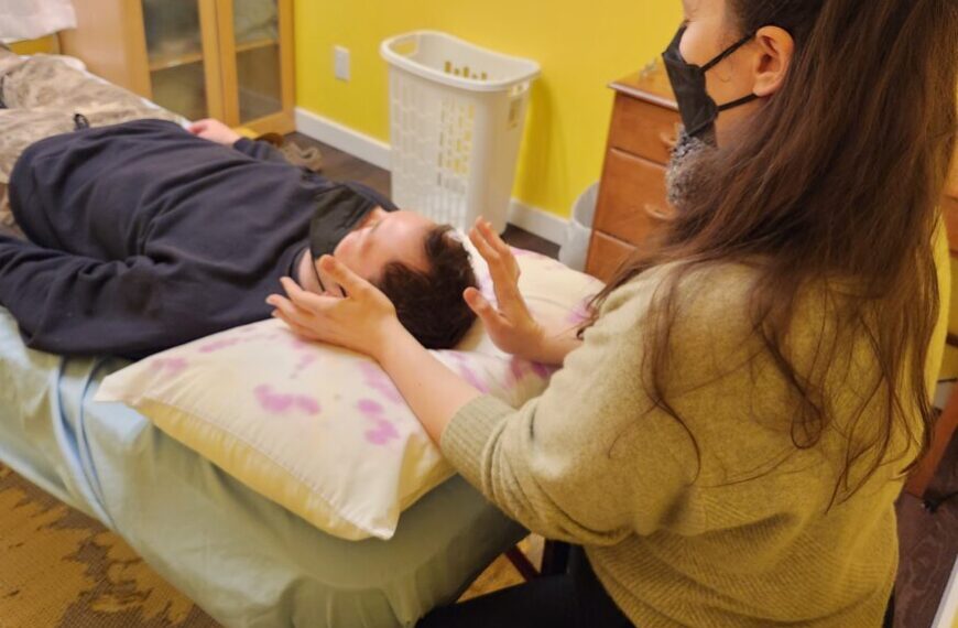 In a room with yellow walls, Alyssa Vincent, wearing a black hoodie, lies on a table. Nearby, another person in a mask gently places their hands near her head, offering Reiki holistic care to create a calming atmosphere.