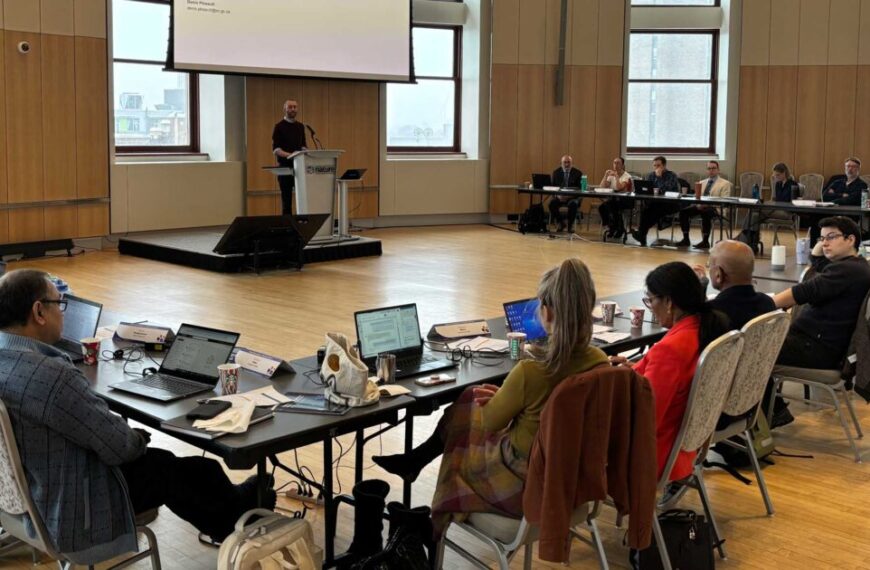 A person speaks at a podium in front of seated attendees around a long conference table in a meeting room. Laptops and papers on the table highlight recent discussions about microplastics. Large windows frame the background, offering a view reminiscent of Canada's vast, open landscapes.