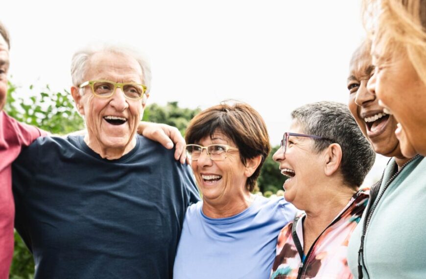 A group of six older adults, emblematic of the silver tsunami, stands close together outdoors, smiling and laughing at the golden opportunity life continues to offer them.
