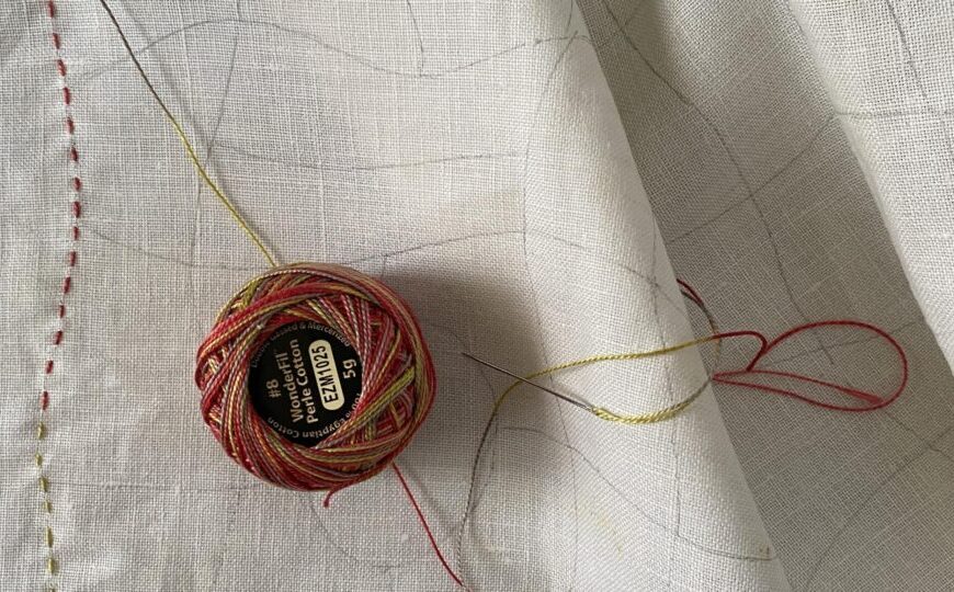 A spool of multicolored thread lies on a white fabric with partially stitched lines, reminiscent of fresh beginnings for the New Year. A row of stitching appears on the left, and the fabric is slightly folded.