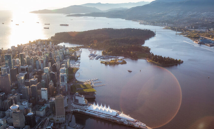 Aerial view of a coastal city with tall buildings alongside a large body of water. A cruise ship is docked at a white-roofed port, surrounded by mountains and forested areas, painting a picturesque story that unfolds with each passing wave.