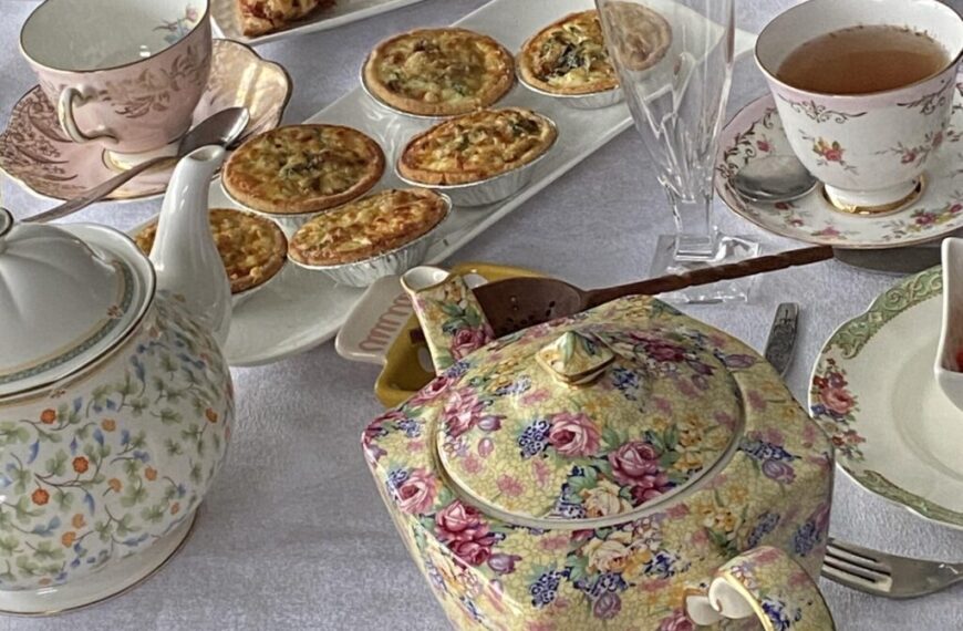 A charming tea setting with floral teapots, teacups, quiche, and pastries graces the white tablecloth. A champagne flute sparkles in the afternoon sun, encapsulating a perfect moment to savor the end of a bustling week.