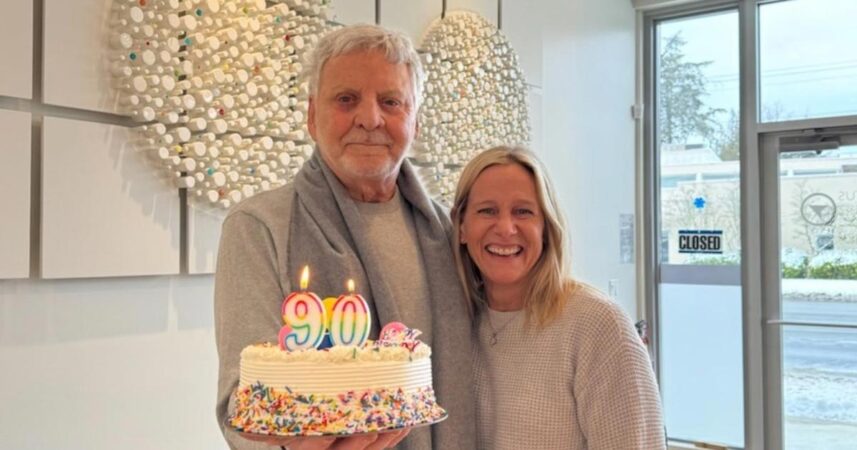 An older man holds a birthday cake with "90" candles, celebrating 90 incredible years, standing beside a smiling woman. They are indoors with decorative wall art in the background.