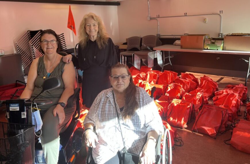 Three women gather in a room filled with red bags scattered on the floor. Two are seated, one in a wheelchair, while the third woman stands behind them. It's a scene of connection and preparation amid tables and stacked chairs in the background.
