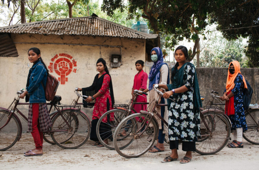 A group of women and girls, perhaps mothers and daughters, stand beside bicycles on a street in front of a wall with graffiti. Some are dressed in vibrant outfits, while one wears a mask. Trees and a building provide the backdrop.