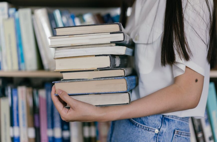 A person balances a stack of books in a library, their white shirt and blue jeans contrasting with the wooden bookshelves. The scene suggests extra time well spent exploring worlds beyond digitally inaccessible readings.