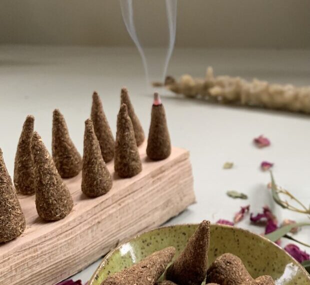 A lineup of incense cones on a wooden holder, one burning and emitting smoke, suggests an enchanting Folk Herbalist's touch. Additional cones rest in a bowl, surrounded by scattered rose petals and dried plants, invoking the ambiance of an Incense Making Workshop.