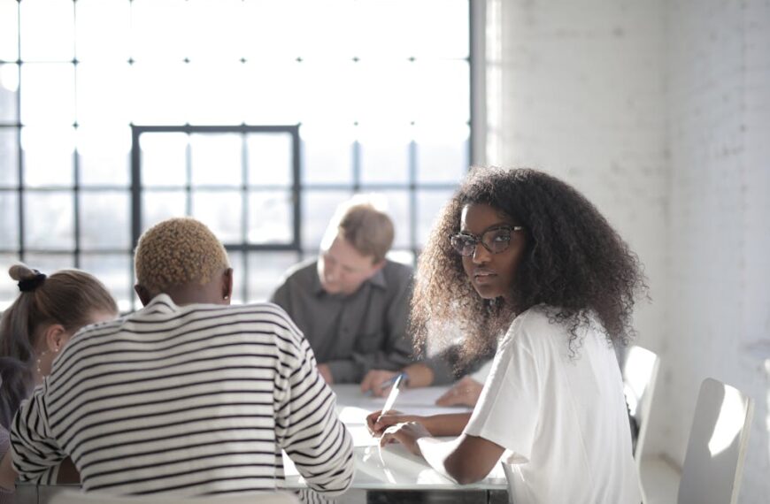 People sitting around a table in a brightly lit room, engaged in discussion. A woman with curly hair and glasses looks towards the camera, seemingly contemplating the technology restrictions mentioned by her colleagues.