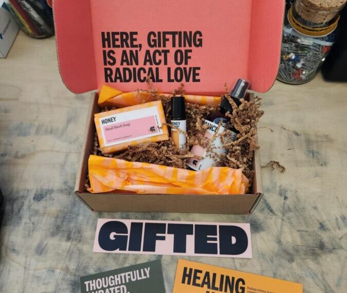 A mysterious package reveals an open gift box filled with skincare treasures, nestled in crinkle paper, with bold text inside the lid. Two cards beside the box proclaim "THOUGHTFULLY CURATED. SURVIVOR MADE." and "HEALING HAPPENS HERE.
