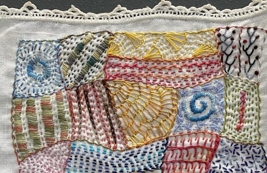 A patchwork embroidery featuring various patterns and colors, including swirls, grids, and wavy lines, already done on a piece of fabric bordered with lace.