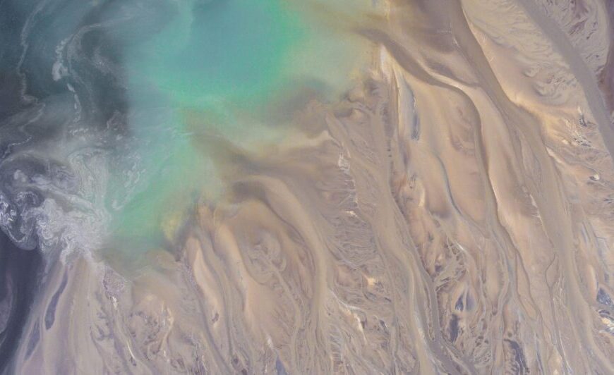 Aerial view of a landscape with swirling turquoise water, beige sand patterns, and light brown sediment forming abstract shapes, inviting Critical Perspectives 2025 on nature’s artistry.