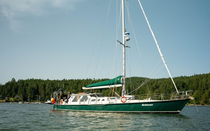 A green sailboat named "Ashmore" floats on calm water near a forested shoreline under a clear sky, as gentle whale vocalizations echo in the distance.