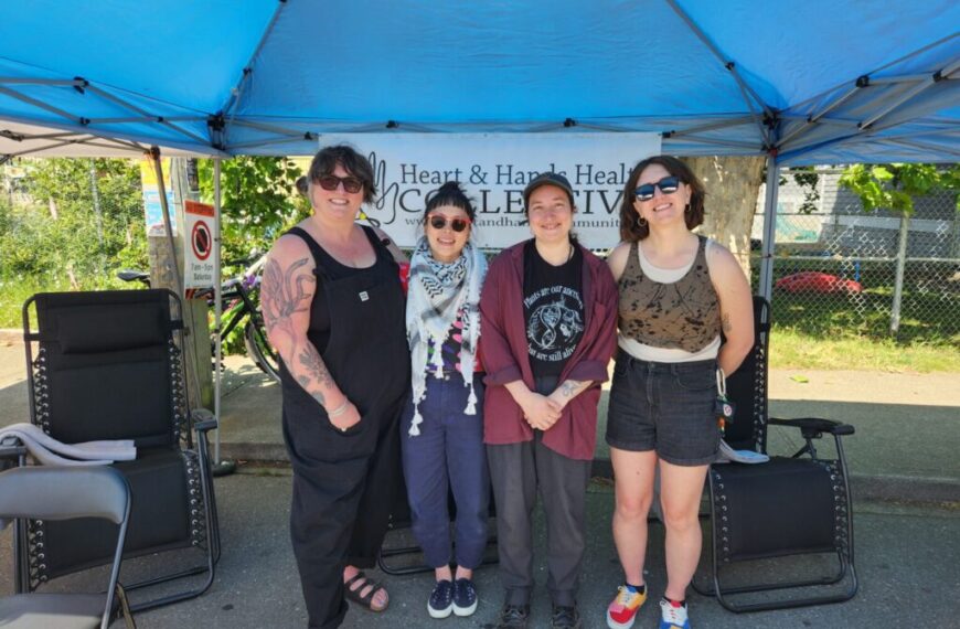 Four people stand and smile under a blue canopy tent with a "Heart & Hands Health Collective" sign behind them, co-creating a welcoming sanctuary in Quadra Village on a sunny day.