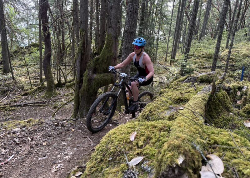 A person wearing a blue helmet and tank top rides a mountain bike on a narrow dirt trail through a forest with moss-covered logs and trees, reminiscent of the XTERRA Victoria course where race results will be tracked in 2025.