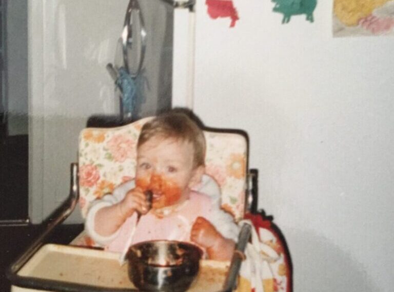 A baby named Bre sits in a high chair eating food with their hands, making a mess. The high chair has a floral pattern, and children’s artwork is displayed on the refrigerator in the background.