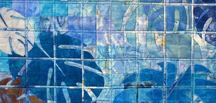 A quilt with blue and white square patches featuring abstract monstera leaf patterns and visible stitching lines, drawing subtle England inspiration.