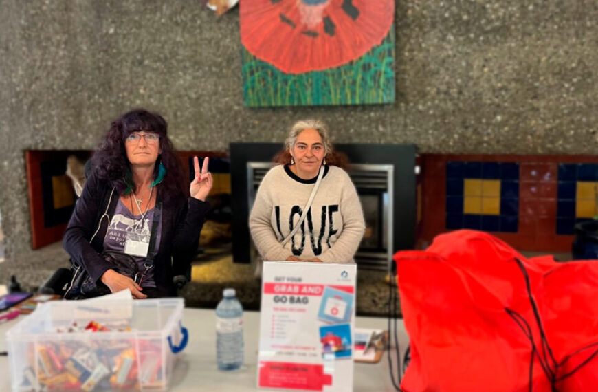 Two women sit behind a table with supplies and red bags for emergencies; one flashes a peace sign. A sign on the table reads "Grab and Go Bag." A large painting hangs behind them.
