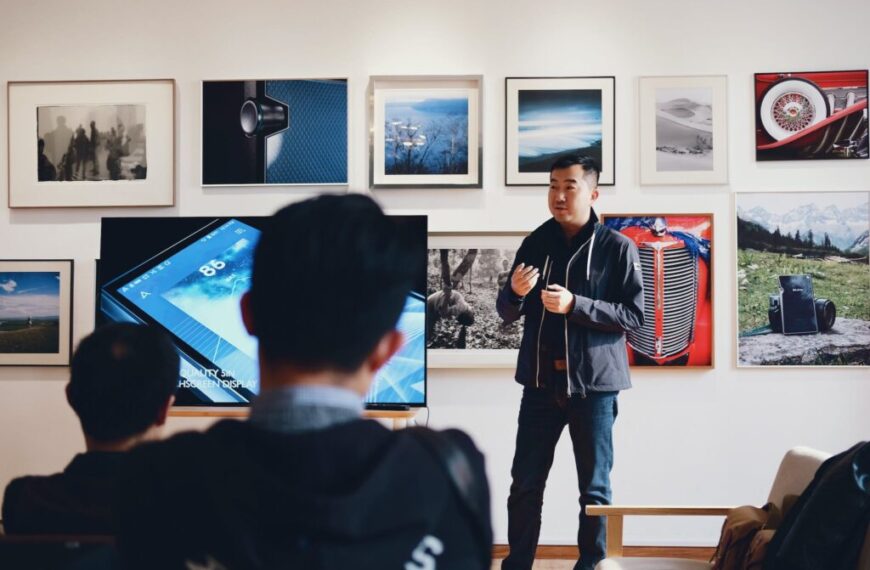 A man stands and leads a presentation assessment to a seated audience in a room with framed photos on the wall and a large screen displaying blue ICT graphics.