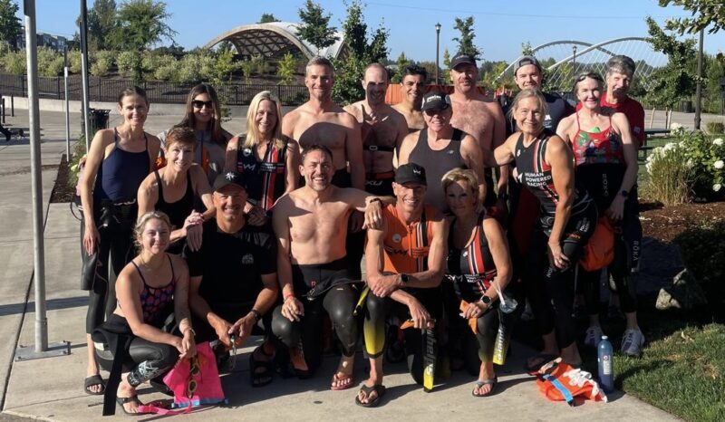 A group of people in wetsuits and athletic gear pose together outdoors on a sunny day, with greenery and a modern bridge structure behind them—capturing the spirit of the Oregon 70.3 for their 2025 race report.