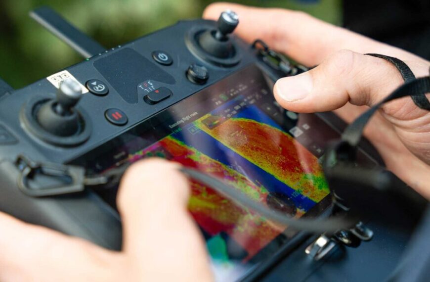 Close-up of hands operating a drone remote control with a screen displaying a colorful aerial thermal image.