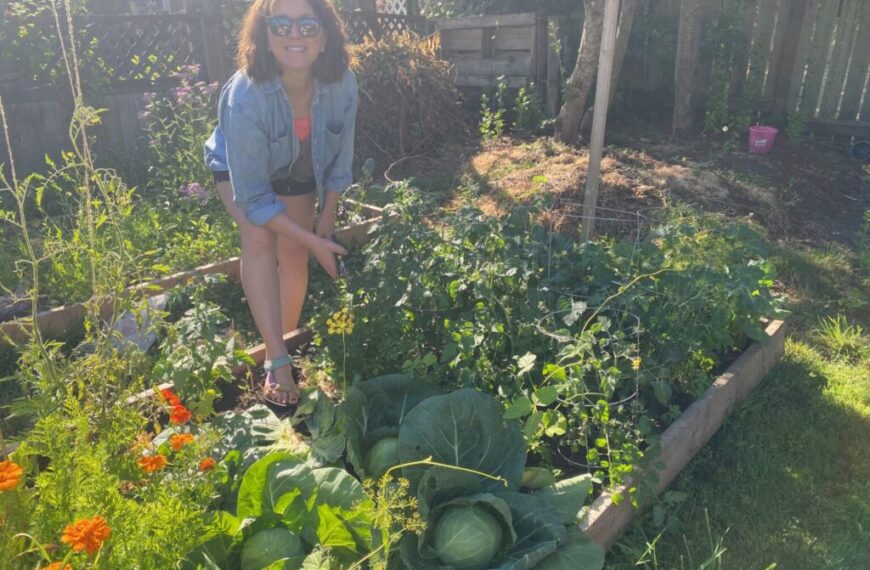 Jade Kulhawy-Bartlett, R.Ac., kneels by a raised garden bed filled with leafy vegetables and flowers, enjoying her journeys in gardening on a sunny backyard afternoon.