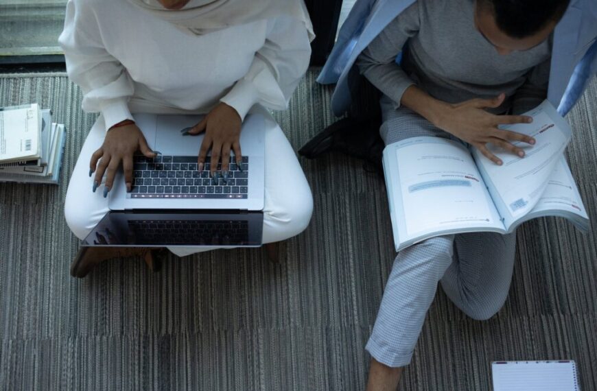 Two learners sit on the floor; one types on a laptop, using ICT to figure out a problem, while the other reads an open book. Books are stacked nearby on the carpet.