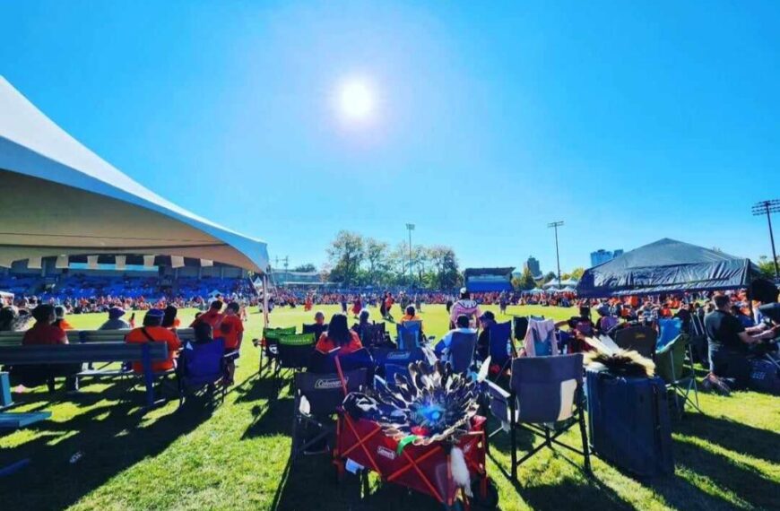 A crowd sits on chairs and under tents at an outdoor stadium event on a sunny day, gathering to observe the National Day of Truth & Reconciliation, with the sun high in a clear blue sky ahead of the Sept 30th 2025 closure.