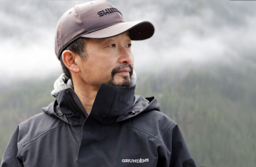 A man wearing a dark jacket and cap stands outdoors, like many anglers, with a misty forest and mountains in the background.