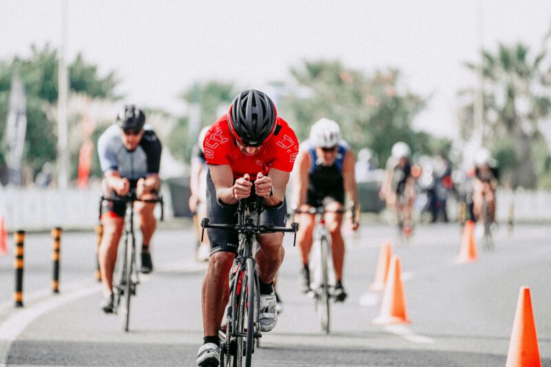 Cyclists participate in a road race, riding in a line on a paved road with orange cones marking the route. Event registration opens October 1st.