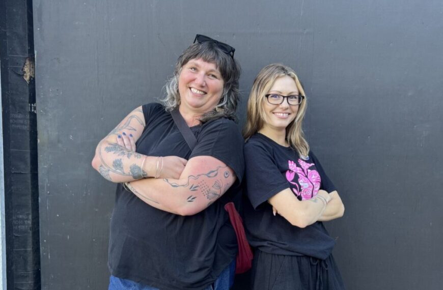 Two smiling women stand back-to-back with arms crossed, both wearing black shirts in front of a plain dark wall—capturing the fun spirit of getting to know each other.