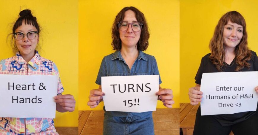 Three people sit against a yellow wall, each holding a sign: "Heart & Hands," "TURNS 15!!," and "Enter our Humans of Heart & Hands anniversary drive <3.