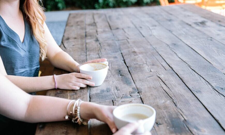 Two people sit at a wooden table holding coffee cups, their arms and torsos in frame, sharing thoughts that will soon be featured in their upcoming podcast.