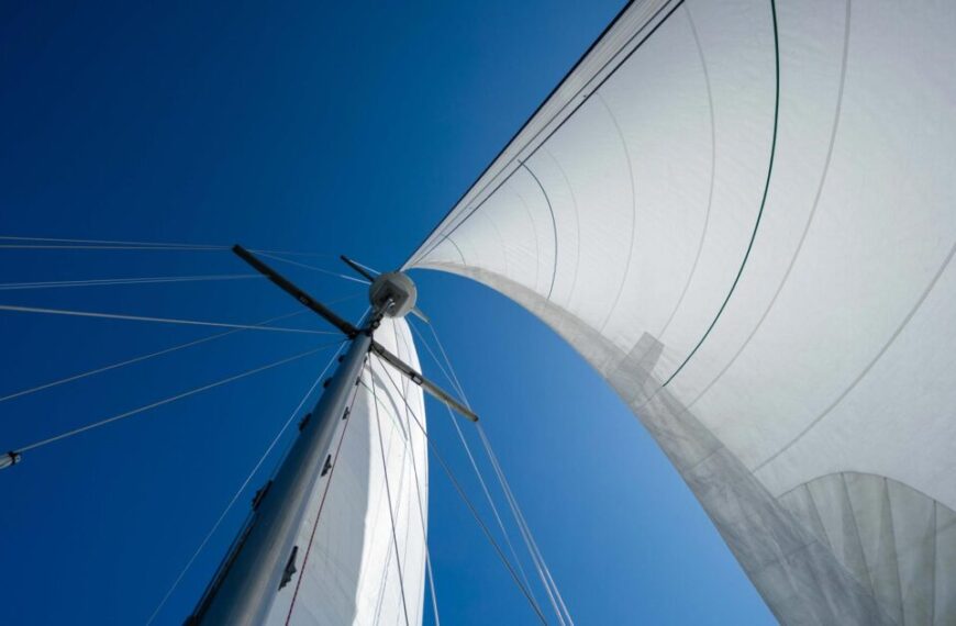 A close-up, upward view of a sailboat mast with white sails unfurled, captured during an educational expedition on the Salish Sea, set against a clear blue sky.