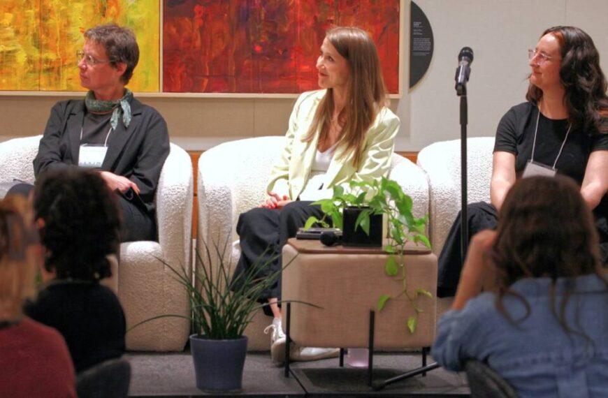 Three women sit on a stage in armchairs, facing left, with plants and a microphone nearby; colorful artwork and a "Living Together 2025" sign decorate the wall behind them.
