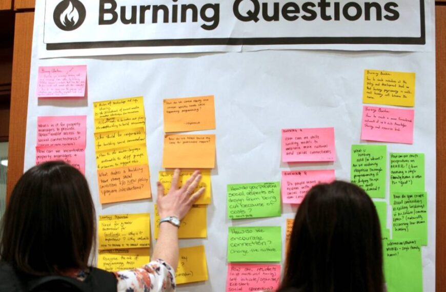 Two people stand in front of a board labeled “Burning Questions,” reading and arranging colorful sticky notes with handwritten questions and comments at the Living Together symposium.