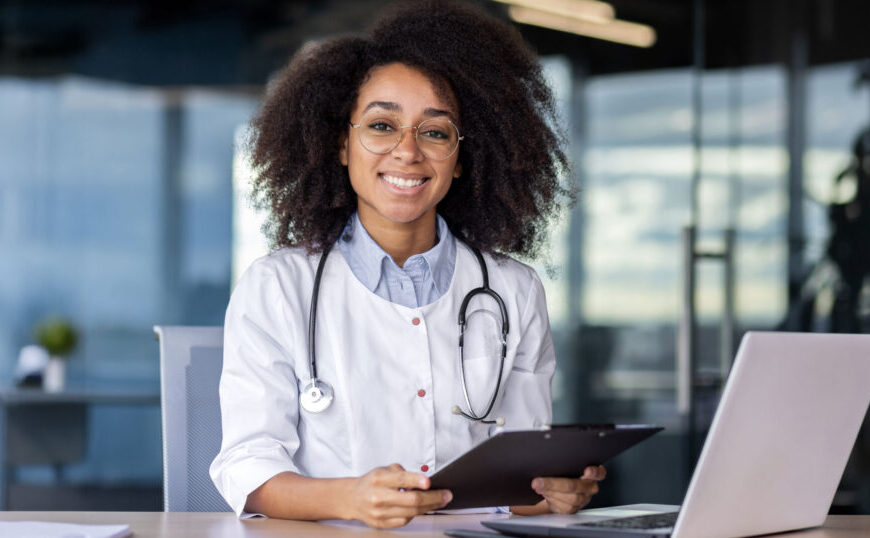 A doctor wearing a white coat and stethoscope sits at a desk with a laptop, holding a clipboard and smiling at the camera, ready to discuss ways to prevent Lyme disease in our health-care system.
