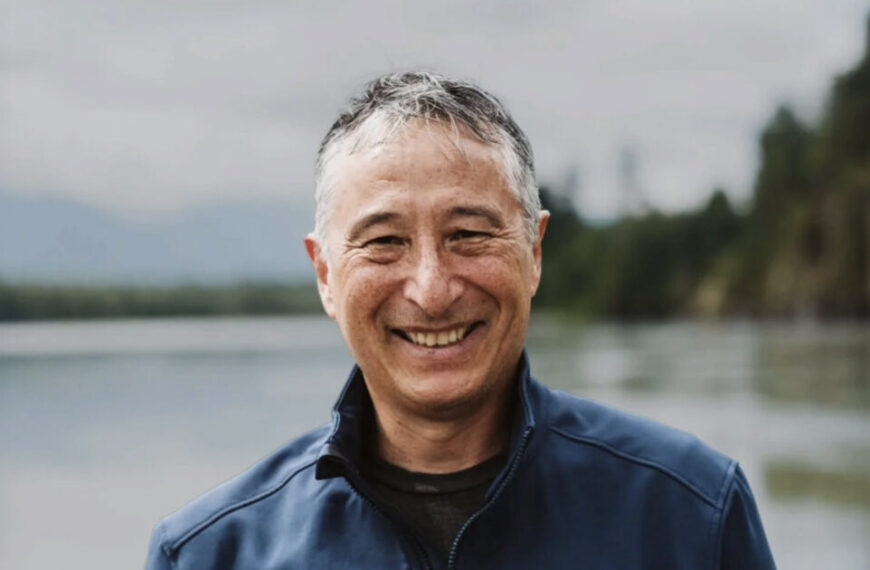 A man wearing a blue jacket smiles outdoors with a blurred lake and trees in the background, reflecting his passion for careers in conservation at organizations like the Pacific Salmon Foundation.