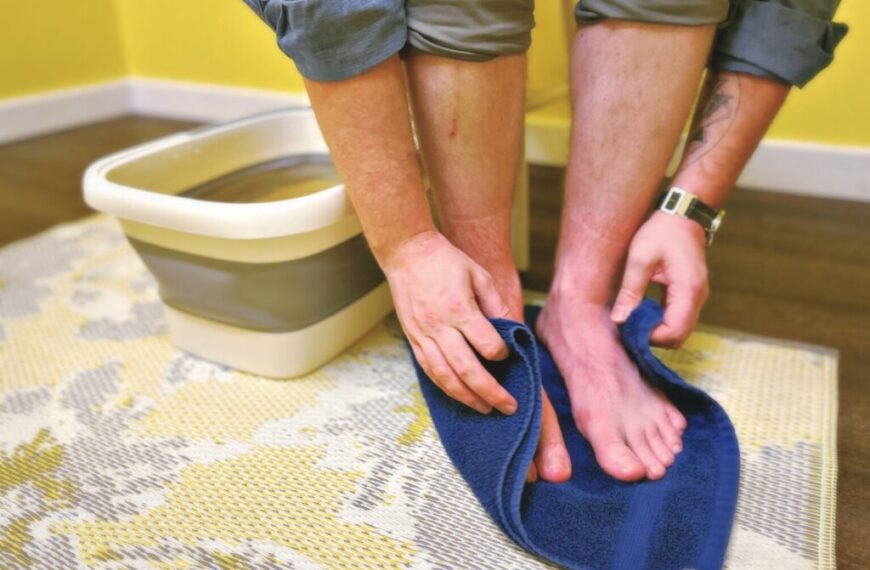 Person drying their feet with a blue towel after a soothing Green Muse Herbs foot soak in a collapsible bath, seated on a chair with rolled-up pants, supporting wellness along the kidney channel.