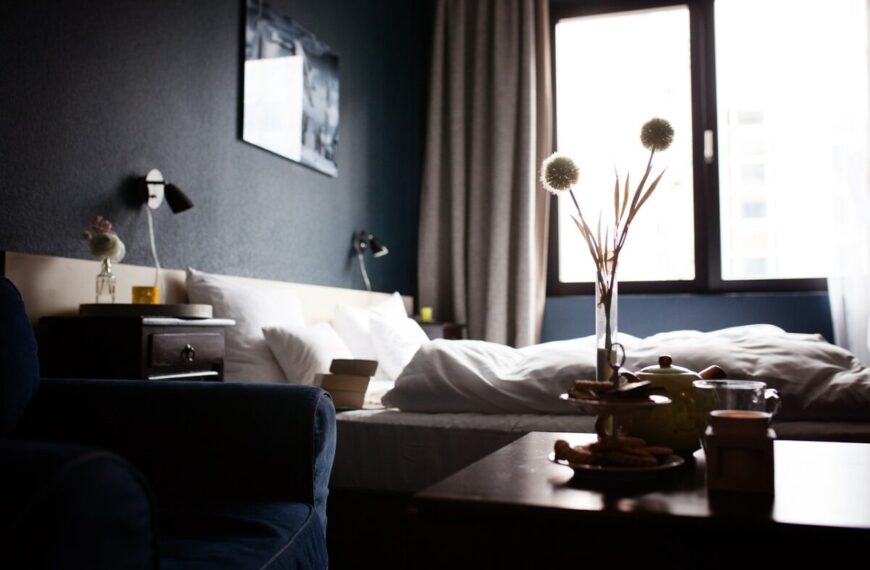 A bedroom with an unmade bed, a nightstand, a dark armchair, and a table set with a teapot, cups, and flowers near a large window with curtains evokes the quiet atmosphere of "Tending The Dead.