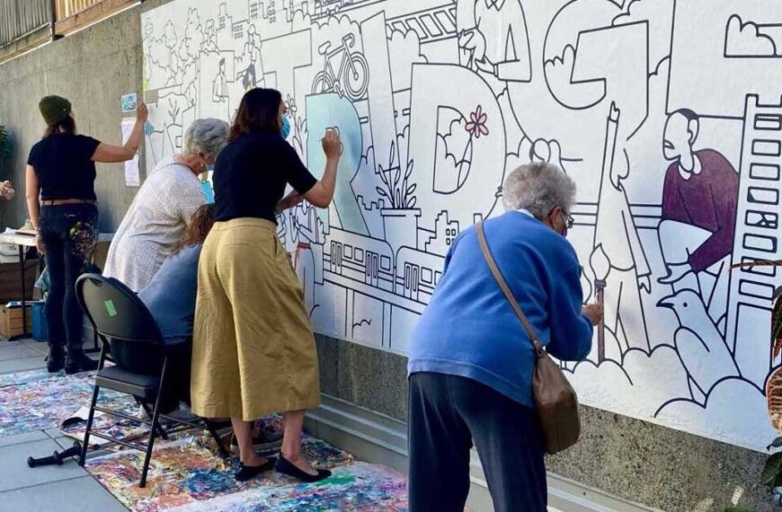 Five people collaboratively paint a large mural outline on an outdoor wall, using brushes and paints, building community as they bring various figures and shapes to life in the mural design.