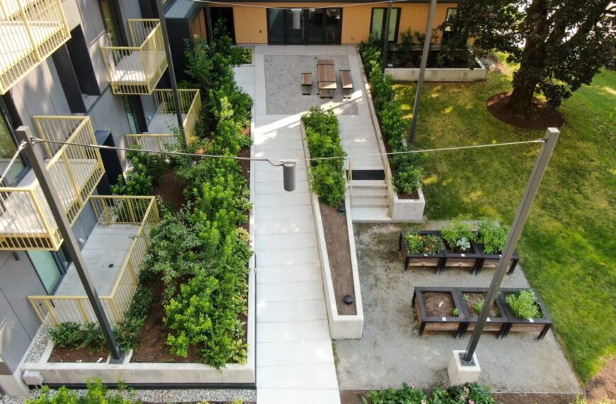 Aerial view of a modern apartment building courtyard by Build Canada Homes, showcasing garden beds, benches, picnic tables, grass, and concrete walkways designed with affordability in mind.